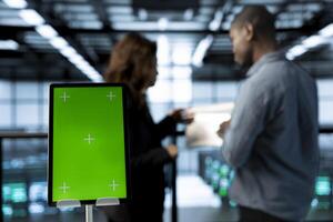 Green screen mockup tablet in front of technicians in blurry background working in server farm, analyzing data. Close up of chroma key tablet next to workers supervising data center photo