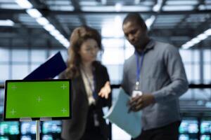 Close up of green screen tablet in front of employees overseeing server room, reading paperwork, troubleshooting hardware. Mockup device next to server hub workers reading technical files photo