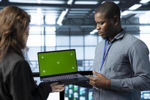 Team of engineers working in server farm, using green screen laptop to analyze data. Employees examining infrastructure in data center using mockup notebook, ensuring system integrity and security photo