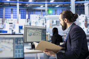 Inspector in solar panel factory checking up on manufacturing equipment, reading documentation files. Man in industrial facility inspecting green energy production process, checking paperwork photo