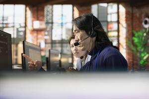 Senior call center agent using headphones microphone to answer customers technical inquiries about hardware issue. Experienced Asian worker doing customer support job using PC to troubleshoot problems photo