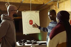 Shooting range safety officer ensuring clients are following rules and operating firearms safely, observing gun holding etiquette. African american firing range gallery worker talking with customers photo