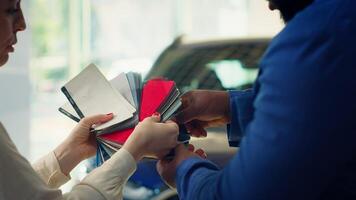 Salesperson in auto showroom discussing with man, offering color swatch samples choice. Agent providing client with color wheel selection, picking hue for car paint, camera A close up photo