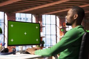 Smiling man enjoying leisure time in apartment, using green screen PC screen to text friends. Happy person talking with mates on social network website using chroma key computer display photo