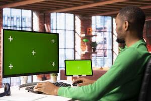 Man in home office using chroma key PC display and tablet to work on job tasks. African american person in apartment workspace using green screen computer monitor and digital device to do job online photo