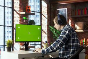 Teleworker in home office listening music and using green screen PC to answer emails from business associates. Remote employee wearing headphones typing on mockup device, solving tasks in apartment photo