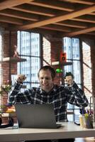 Happy man at home office desk celebrating after seeing he achieved sales target on laptop screen. Cheerful employee excited after receiving promotion at work, reading email on notebook photo