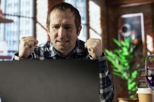 Cheerful worker at home office desk celebrates after seeing he achieved sales target on laptop screen. Close up of jolly employee delighted after receiving promotion at work, reading email on notebook photo