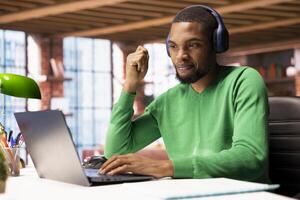Remote employee wearing headphones at home participating in online training session on laptop. Teleworker using audio device at home to watch instructional course videos on notebook photo