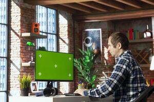 Person at home office desk browsing internet on green screen computer monitor, researching information online. Man coding software at workspace desk, writing and testing program code on chroma key PC photo