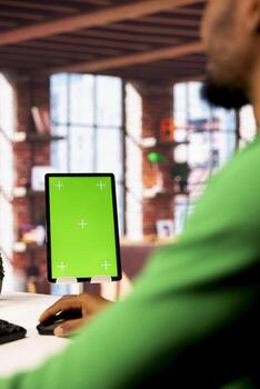 Green screen tablet on desk next to man in home office typing on PC keyboard. Mockup device next to person in apartment reading articles on PC screen, gathering data for work project photo
