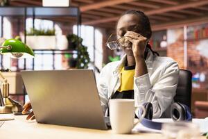 Overworked woman in apartment removing eyeglasses, trying to relax eyes after working too much on laptop. Remote worker feeling eye strain, getting overstimulated while working from home photo