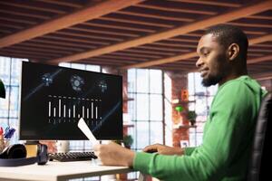 Smiling man in home office using financial application UI on PC screen showing company revenue and expenses. Accountant working from apartment doing annual profits review using computer display photo