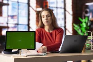 Freelancer does research by reading articles and papers next to chroma key display, collecting information for new project. Woman multitasking and studying files with handwritten notes at the desk. photo