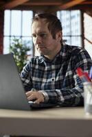 Man at home office desk edits documents on laptop, updating reports and files, typing text, formatting paragraphs, inserting images, saving changes, managing digital documents within file directories photo