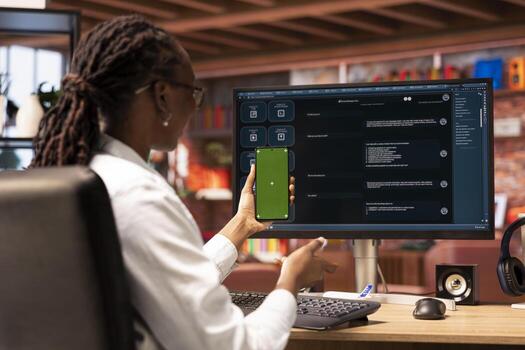 Happy woman uses green screen phone, doing videocall in front of PC with AI chatbot answering prompt. Cheerful african american person uses LLM program during videoconference on mockup smartphone photo