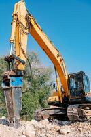 Yellow drilling machine drills stones in a quarry while standing near a green tree photo