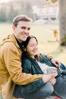 Smiling man hugging woman with mug sitting in autumn park photo