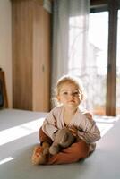 Little girl hugging a teddy bear while sitting on the bed in the bedroom and leaning forward photo
