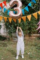 Little girl stands on a green lawn under colorful flags on a string and holds an inflatable number 3 photo