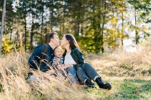 Dad and mom kiss while sitting with a little girl in a clearing near the forest photo