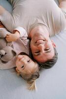 Smiling dad with a little girl and a teddy bear lie on the bed touching their heads. Top view photo
