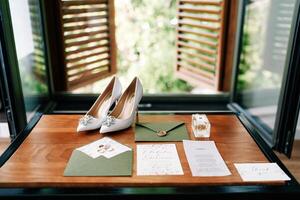 Bride shoes stand on a wooden table next to invitations in green envelopes near the window photo