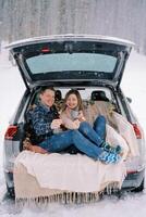 Smiling boyfriend and girlfriend are sitting in the trunk of a car under snowfall with sparklers and mugs of coffee photo
