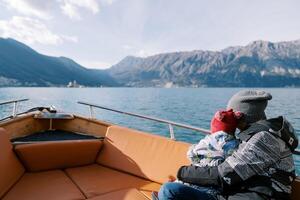 Mom with a small child in her arms floats in a boat along sea and looks at the mountains. Back view photo