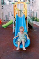 Little smiling girl sitting at the bottom of a plastic slide on a playground in the yard photo