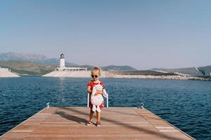 Little girl with a pink toy rabbit stands on the pier against the background of a lighthouse on the shore photo