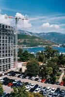Montenegro, Budva - June 12, 2024. Parking lot with cars in front of a tall building under construction in scaffolding near a tower crane photo