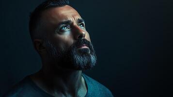 Bearded man gazes upwards with thoughtful expression intense gaze against dark background Light shines on face photo