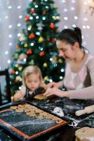 Tray of cut-out cookies stands on a table next to a mother and little daughter making cut-outs photo