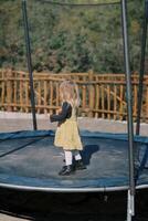 Little girl stands on a trampoline with a mesh fence in a green park. Side view photo