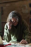 Man talking on smartphone and typing on laptop in workshop photo