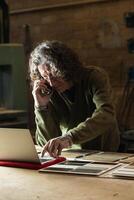 Man talking on smartphone and typing on laptop in workshop photo