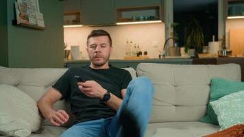 Man sits on sofa, holding TV remote control in hand. Person relaxing on couch and watching television. Concept of relaxation, leisure time and home entertainment video