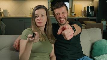 Smiling couple sitting on sofa watching TV together at home. Man and woman laughing while watching television in living room. Concept of relaxation, entertainment and spending time together video