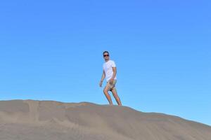 A young man walking along a sand dune through barkhan in Maspalomas, Gran Canaria on a hot day. A self confident man goes up the mountain - success and achievements concept photo