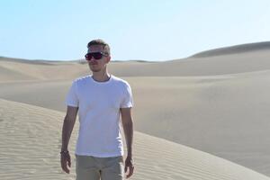 A stylish model portrait of a young man walking along a sand dune through barkhan in Maspalomas, Gran Canaria on a hot sunny day. Desert travel touristic concept photo
