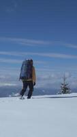 A person wearing a yellow jacket walks through deep snow in the mountains, with a large backpack. Surrounding snow-capped peaks contrast against a bright blue sky. Vertical footage video