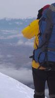 A traveler in a yellow jacket with a backpack climbs a snowy peak among the clouds, enjoying the view. Vertical footage video