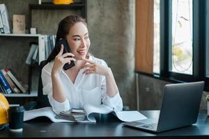 Businesswoman multitasking in a modern office, talking on the phone, using a laptop, and reviewing documents. Bright and productive workspace. photo