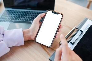 Close-up of hands holding a smartphone with a blank screen, next to a laptop and clipboard on a wooden desk. photo