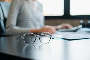 Modern eyeglasses on a desk with a blurred person working on a computer, symbolizing focus and productivity. photo
