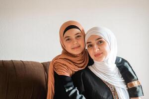 Happy Muslim family at home young mother and child daughter in hijab hug with love on sofa together for bonding with affection on mothers day. photo