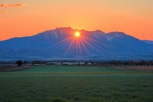 Sun Setting Behind A Mountain Range With Sunbeams Over Field photo