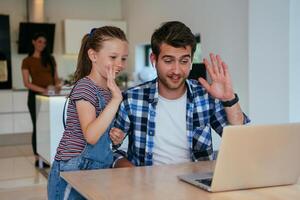 Work from home. Daughter interrupting her father while he is having a business online conversation on his laptop while sitting in the modern living room. photo