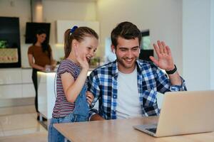 Work from home. Daughter interrupting her father while he is having a business online conversation on his laptop while sitting in the modern living room. photo
